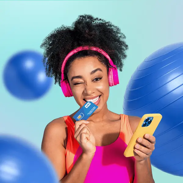 Woman in pink headphones biting a Tello SIM card while holding her phone, surrounded by exercise balls